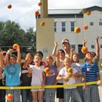 group of young kids standing behind tennis net throwing tennis balls in the air